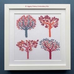 four different colored trees are displayed in a white frame on a blue wall, with the words happy pekay embroiderry kits written below them