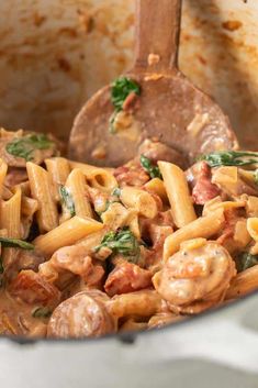 a pot full of pasta with meat and spinach on the side next to a wooden spoon