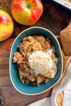 an apple crisp with ice cream in a blue bowl next to two apples on a wooden table