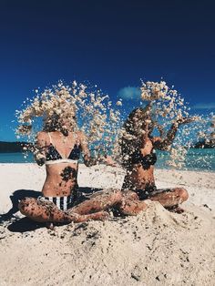 two women in bathing suits splashing water on each other while sitting on the beach