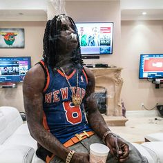 a man with dreadlocks sitting on a couch in front of two televisions