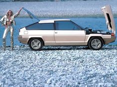 a woman standing next to a car with its hood open on the beach near water