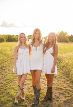 three beautiful young women standing next to each other in a field wearing cowboy boots and dresses