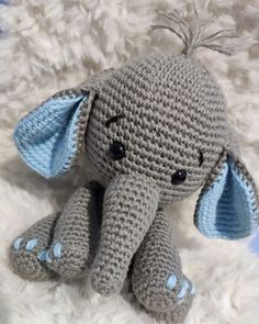 a crocheted gray elephant sitting on top of a white blanket with blue ears