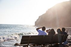 four people sitting on a bench near the water and rocks at the beach, looking out to sea