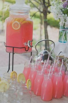 a table topped with lots of glasses filled with pink liquid and lemons next to a beverage dispenser