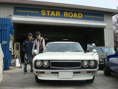 two people standing in front of a star road garage with their cars parked outside it