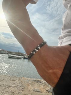 a man wearing a bracelet on his wrist next to the water with boats in the background