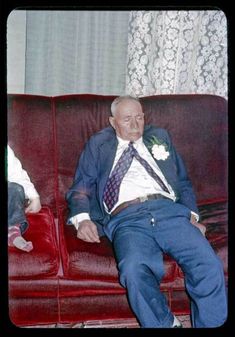 an old man wearing a suit and tie sitting on a red couch next to another older man