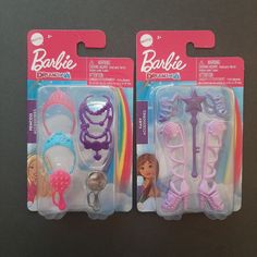 two barbie dolls are in plastic packaging on a black table with pink and purple hair accessories