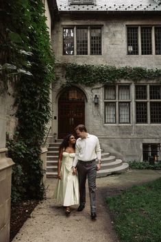 a man and woman walking down a path in front of a building with ivy growing on it