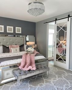 a bedroom decorated in gray and pink with a bed, mirror closets, chandelier, and ottoman