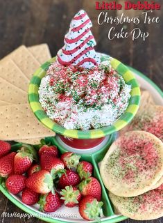 a christmas cake dip surrounded by crackers and strawberries on a green plate with the words, little debite christmas tree cake dip