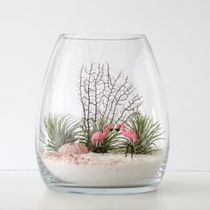 two pink flamingos in a glass vase filled with sand and plants