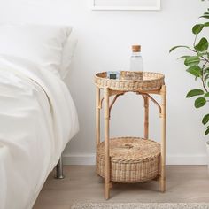 a wicker table next to a bed with a bottle on top and a plant in the corner