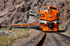 an orange train traveling down the tracks next to a mountain side covered in grass and rocks