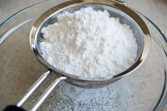 a metal strainer filled with white powder on top of a glass table next to a measuring spoon