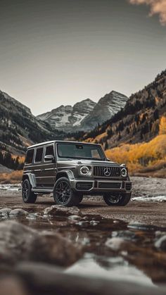 a jeep parked on the side of a river in front of mountains and snow capped peaks