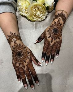 two hands with henna tattoos on them and flowers in the background, one is holding a