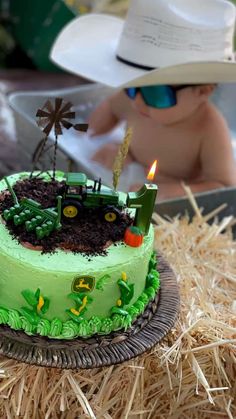 a green cake with a tractor on it sitting in hay next to a baby wearing a cowboy hat