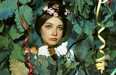 a woman with flowers in her hair is surrounded by green leaves and ribbons as she looks into the camera