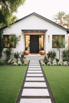 a white house with grass and plants on the front lawn, leading to a black door