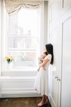 a woman in a white dress holding a baby next to a window with flowers on the windowsill