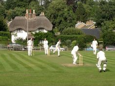 a group of men playing a game of cricket in front of a white thatched house