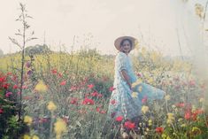 a woman standing in a field full of wildflowers wearing a hat and dress