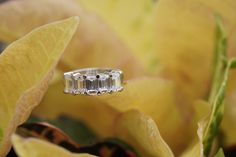 a three stone diamond ring sitting on top of a leafy green plant with yellow flowers in the background