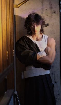 a man with his arms crossed standing in front of a wall wearing a white shirt and black skirt