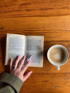 a person's hand on top of an open book next to a cup of coffee