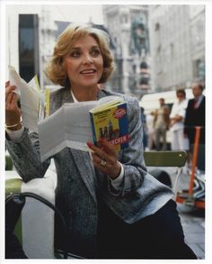 a woman sitting down reading a book and holding something in her hand while looking at the camera