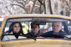 three people are sitting in the driver's seat of a yellow car on a city street