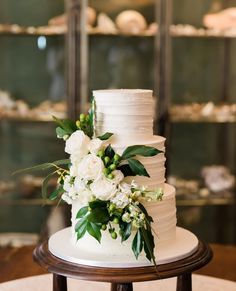 a wedding cake with white flowers and greenery sits on a table in front of glass cases