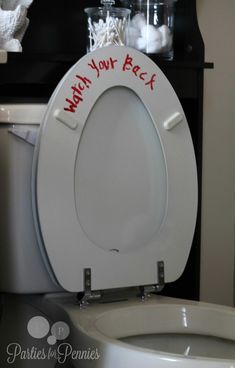 a white toilet with red writing on the lid next to a trash can and shelf