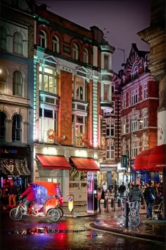 people are walking down the street in front of some buildings at night with lights on