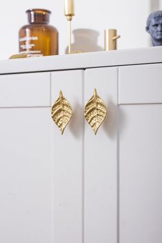 a pair of gold leaf shaped earrings sitting on top of a white cabinet next to a vase