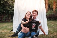 a man and woman sitting on the grass holding up a t - shirt