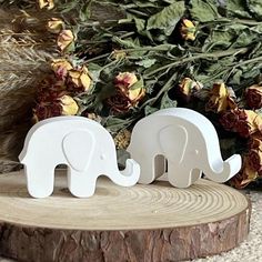 two white elephant figurines sitting on top of a tree stump next to flowers