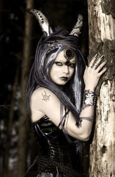 a woman with long black hair and piercings on her head leaning against a tree