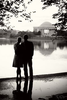 two people standing next to each other in front of a lake