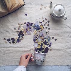 a person holding a coffee cup with flowers on the table next to an open book