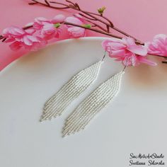 the earrings are made with pearls and beaded in silver color, sitting on a white plate next to pink flowers