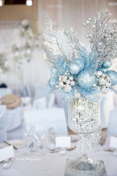 a vase filled with blue and silver ornaments on top of a table covered in white linens