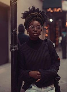 a woman standing next to a pole with her arms crossed wearing glasses and a turban