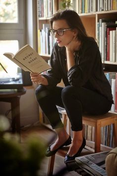 a woman sitting on a chair reading a book in front of a bookshelf