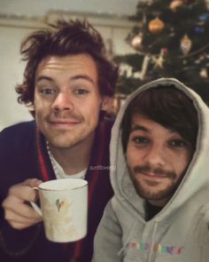 two young men are posing for the camera while holding coffee cups in front of a christmas tree