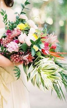 a woman holding a bouquet of flowers and greenery in her hands while wearing a white dress