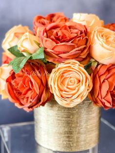 an arrangement of orange and yellow roses in a gold vase on a clear table top